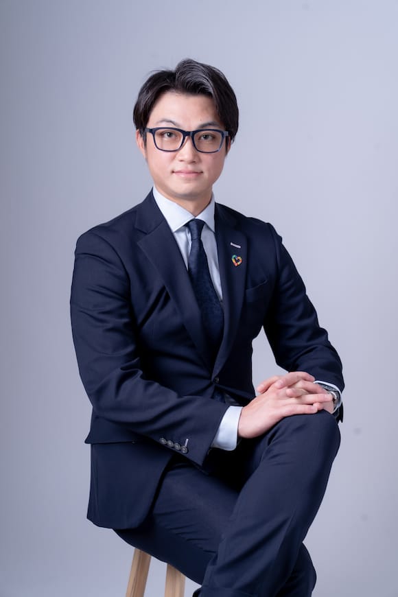 淡いグレー背景でネイビースーツの着座ポートレート、会社HP用のビジネスプロフィール写真